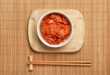 Photo of Bowl of spicy cabbage kimchi and chopsticks on bamboo mat, flat lay