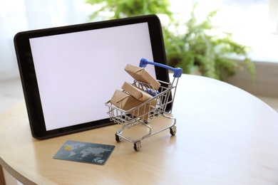 Internet shopping. Small cart with boxes and credit card near modern tablet on table indoors