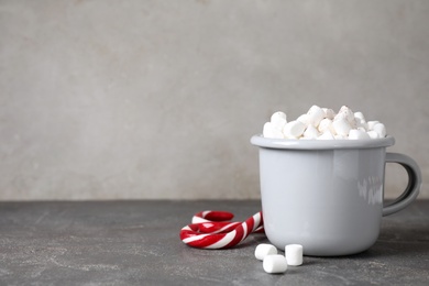 Cup of tasty hot drink with marshmallows near candy canes on grey table. Space for text