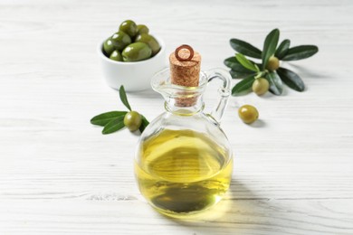 Glass jug of oil, ripe olives and green leaves on white wooden table