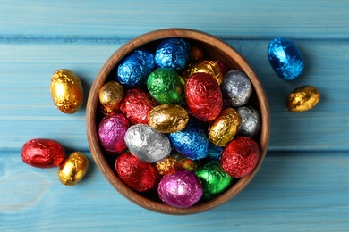 Bowl with chocolate eggs wrapped in colorful foil on light blue wooden table, flat lay