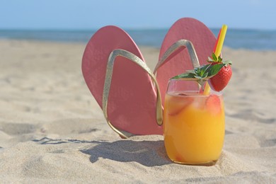 Photo of Glass of refreshing drink with strawberry and flip flops on sandy beach near sea