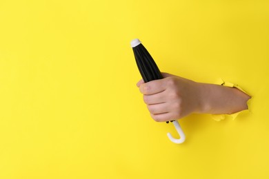 Woman holding closed small black umbrella through hole in yellow paper, closeup. Space for text