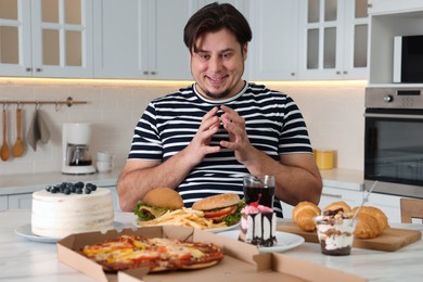 Photo of Hungry overweight man at table with sweets and fast food in kitchen