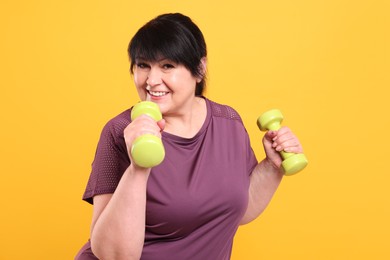 Happy overweight mature woman doing exercise with dumbbells on orange background