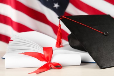 Black graduation hat, diploma and book on white wooden table against American flag