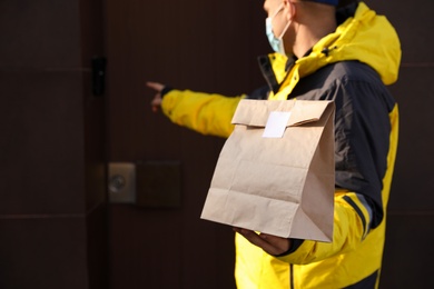 Courier in medical mask ringing doorbell outdoors, focus on paper bag with takeaway food. Delivery service during quarantine due to Covid-19 outbreak
