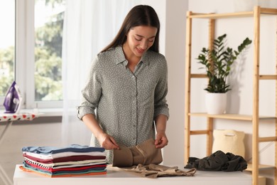 Photo of Young woman folding clothes at white table indoors