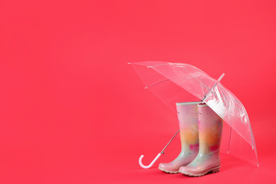 Transparent umbrella and colorful rubber boots on red background. Space for text