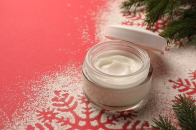 Photo of Winter skin care. Hand cream near snowflake silhouettes made with artificial snow and fir branches on red background, closeup