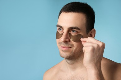 Young man applying under eye patches on light blue background. Space for text