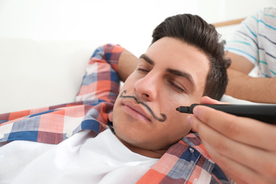 Photo of Young man drawing mustache on face of sleeping friend, closeup. April fool's day