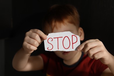 Photo of Little boy holding piece of paper with word Stop against black background, focus on hands. Domestic violence concept