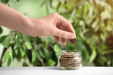 Woman putting coin into jar on white wooden table against blurred green background, closeup. Money savings