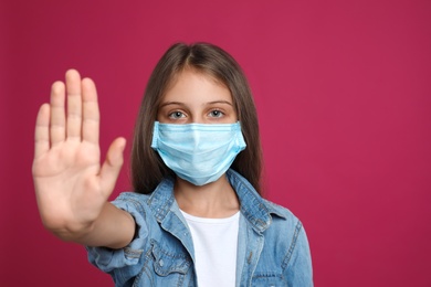 Little girl in protective mask showing stop gesture on crimson background, space for text. Prevent spreading of coronavirus