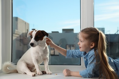 Cute little girl with her dog on window sill indoors. Childhood pet