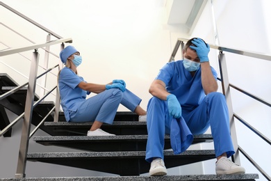Exhausted doctors sitting on stairs indoors. Stress of health care workers during COVID-19 pandemic