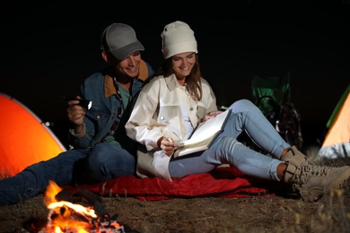 Couple with flashlight reading book near bonfire at night. Camping season
