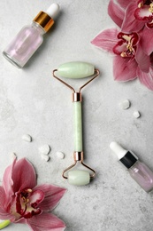 Natural jade face roller, cosmetic product and flowers on grey background, flat lay