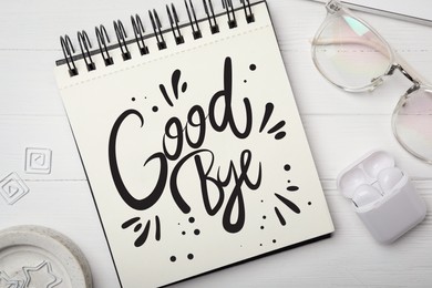 Notebook with word Goodbye, eyeglasses and earphones on white wooden table, flat lay