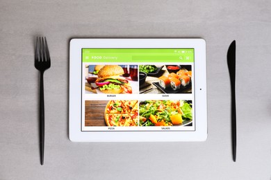 Modern tablet with open page for online food ordering and cutlery on grey table, flat lay. Concept of delivery service