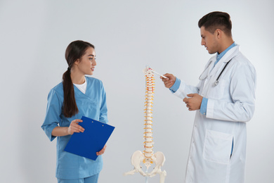 Professional orthopedist with human spine model teaching medical student against light background
