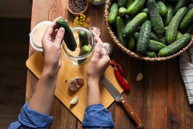 Woman putting cucumbers into jar at wooden table, top view. Pickling vegetables