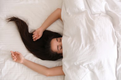 Young woman hiding under warm white blanket in bed, top view