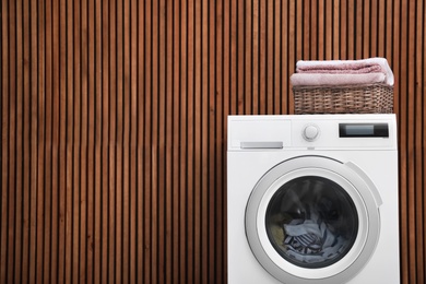 Washing machine and laundry basket with towels near wooden wall. Space for text