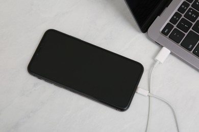 Smartphone connected with charge cable to laptop on light table, above view. Space for text