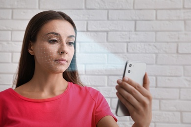 Young woman unlocking smartphone with facial scanner near white brick wall. Biometric verification