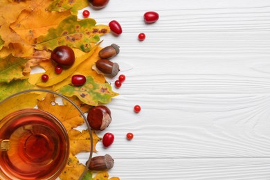 Photo of Different autumn leaves, acorns, chestnuts, red berries and cup of tasty tea on white wooden table, flat lay. Space for text
