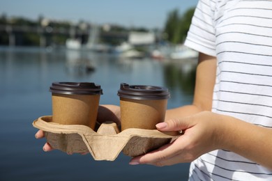 Woman holding takeaway paper coffee cups with plastic lids in cardboard holder near river, closeup