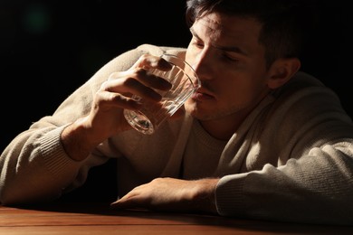 Addicted man drinking alcohol at wooden table against black background