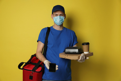 Photo of Courier in protective mask holding order and smartphone for contactless payment on yellow background. Food delivery service during coronavirus quarantine