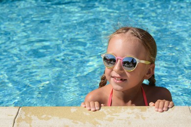 Happy cute little girl with sunglasses at edge of swimming pool on sunny day. Space for text