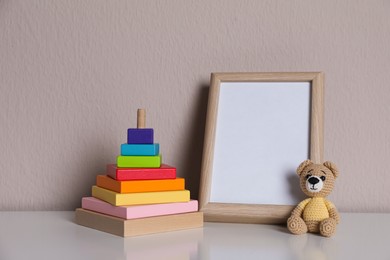 Empty photo frame, toy bear and pyramid on white table near grey wall. Space for design