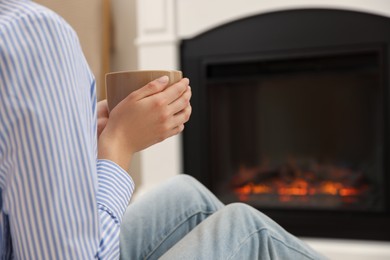 Young woman with cup of hot drink resting near fireplace at home, closeup