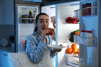 Young woman eating bun near open refrigerator at night