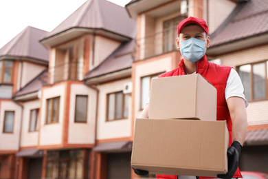 Courier in protective mask and gloves with boxes near house outdoors. Delivery service during coronavirus quarantine