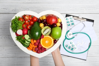 Female doctor holding plate with fresh fruits and vegetables over table, top view. Cardiac diet