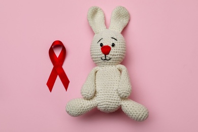 Photo of Cute knitted toy bunny and red ribbon on pink background, flat lay. AIDS disease awareness