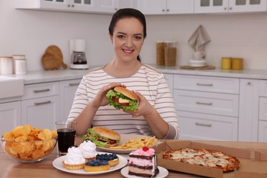 Happy overweight woman with burger and other unhealthy food in kitchen