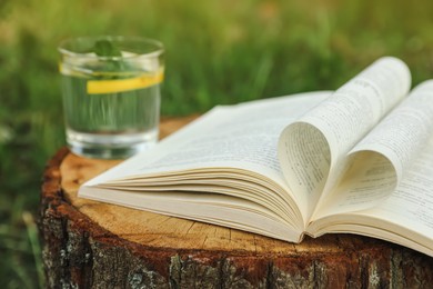 Open book near glass of water with mint and lemon on tree stump outdoors, closeup