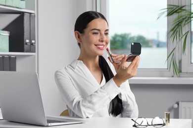 Photo of Young woman using cosmetic pocket mirror while applying makeup in office