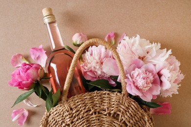 Wicker bag with bottle of rose wine and beautiful pink peonies on brown background, top view