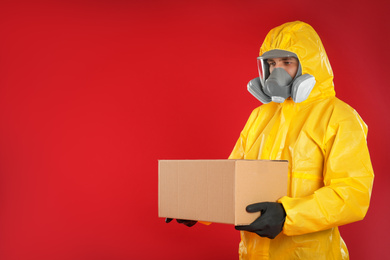 Man wearing chemical protective suit with cardboard box on red background, space for text. Prevention of virus spread