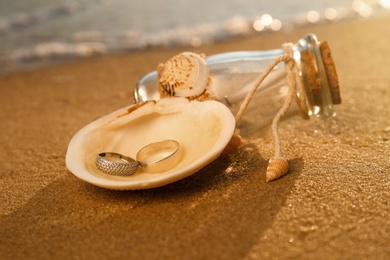 Shell with gold wedding rings and invitation in glass bottle on sandy beach at sunset, closeup
