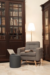 Comfortable armchair with pouf, laptop and lamp near wooden bookcase in library