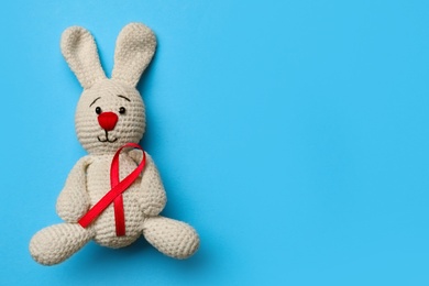 Photo of Cute knitted toy bunny with red ribbon on blue background, top view and space for text. AIDS disease awareness